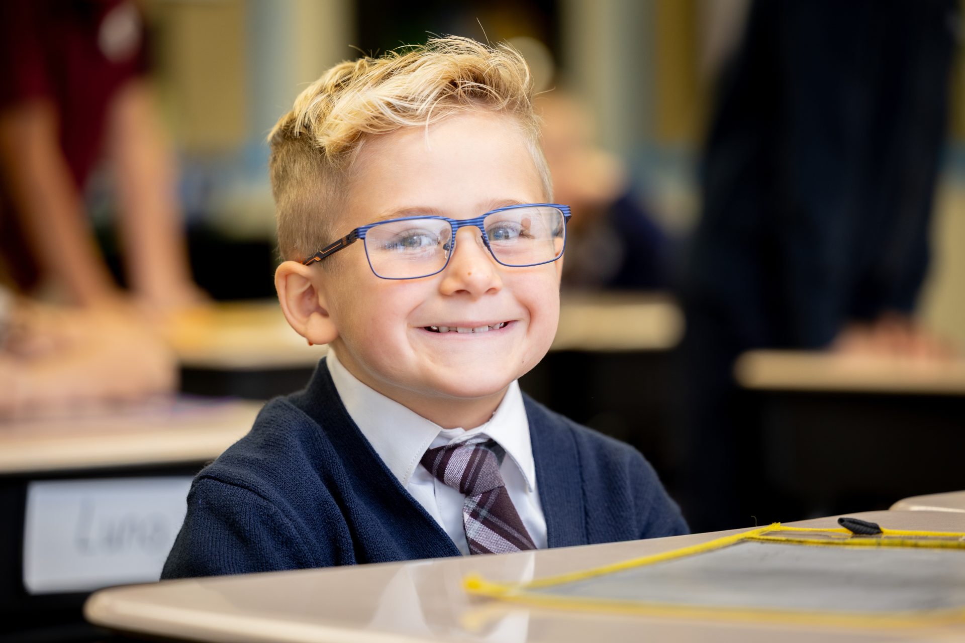 Cheerful boy in a classroom wearing glasses, blue cardigan, and smiling with joy.