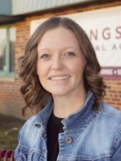 Friendly woman in a denim jacket smiles warmly against a rustic brick wall background.